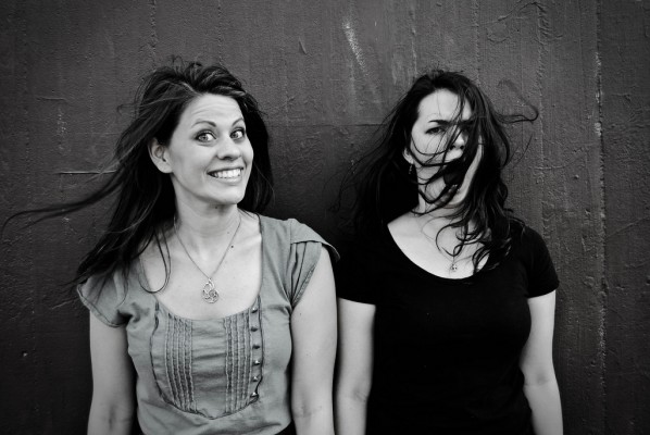 black and white photo of two women against a wall. One is smiling and looking at the camera, the other is yawning with the wind blowing her long, dark hair into her face