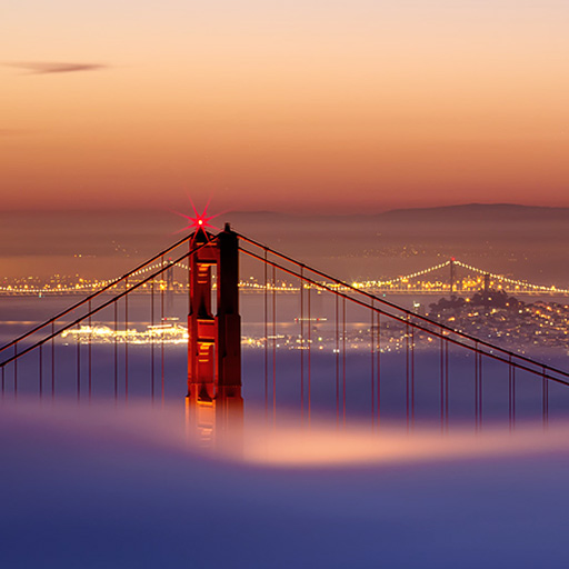 photo of fog shrouded Golden Gate Bridge with an orange glow in the distance and dawn on the horizon