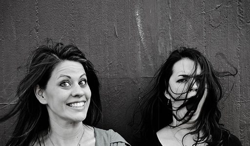 black-and-white photo of 2 women standing side by side and against a wall. One has a big smile when the wind blows the other's hair.